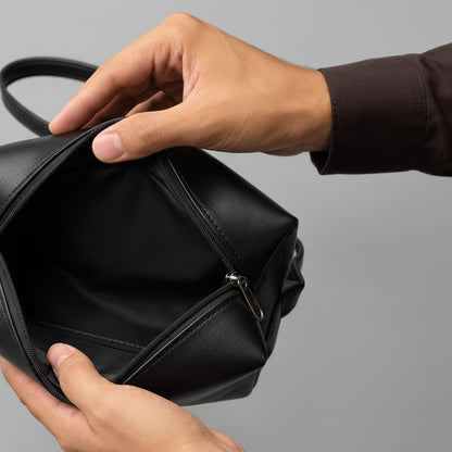 A close-up of a person's hands holding open a tan leather bag to show its black interior lining, set against a solid gray background.