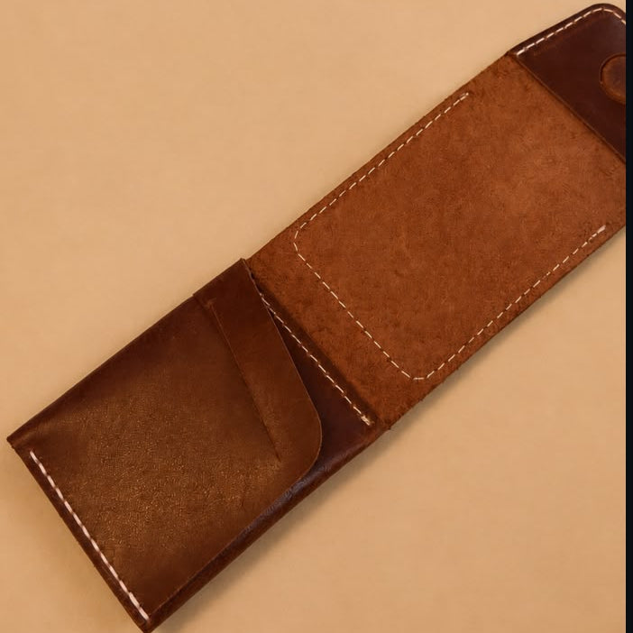 A brown leather sunglasses case with white contrast stitching, shown open on a warm beige background with a pair of black sunglasses placed in the upper right corner.