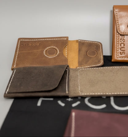 A brown leather card holder wallet with a circular magnetic closure and contrast stitching, displayed open on a wooden surface with a black branded dust bag underneath.