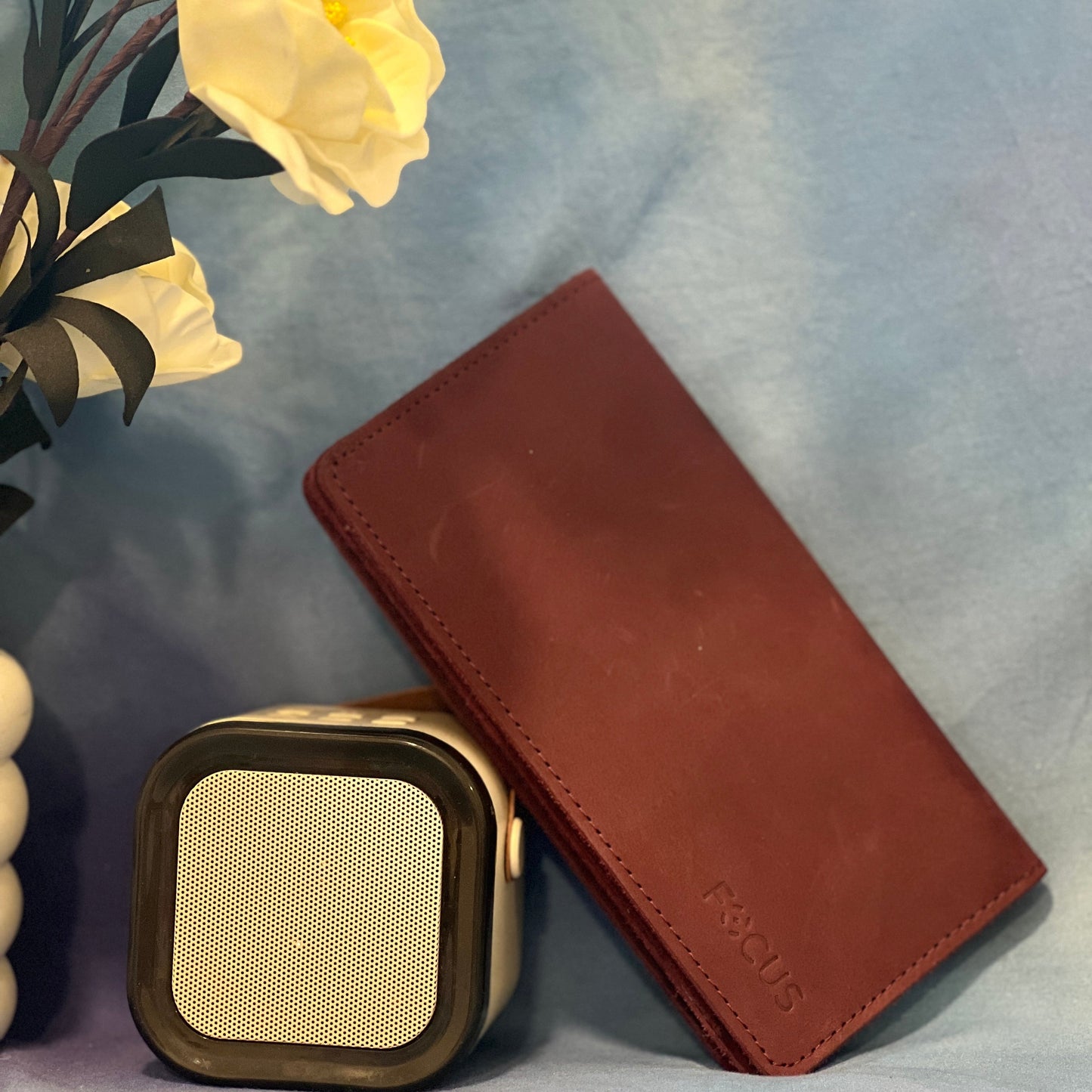 Brown leather wallet and square gold object on a light blue surface with yellow flowers.