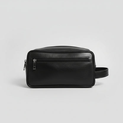 A black faux-leather rectangular pouch bag with a front zippered pocket and a side handle, resting on a glass table in an indoor setting.