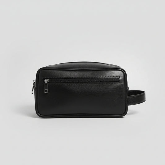 A black faux-leather rectangular pouch bag with a front zippered pocket and a side handle, resting on a glass table in an indoor setting.