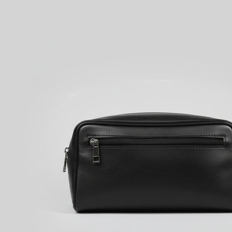 A black leather zippered pouch or small bag with a silver zipper pull, photographed on a reflective glass tabletop.